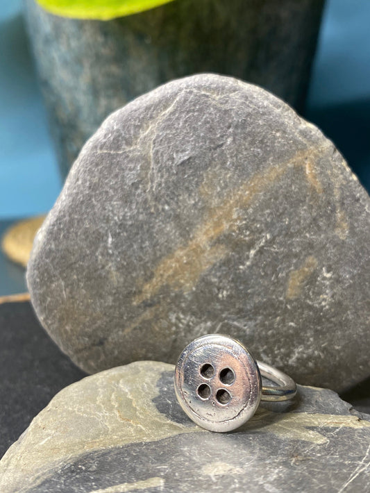 Silver ring with four holes on a stone surface with green leaves in the background.
Inspired by the Borrowers novel