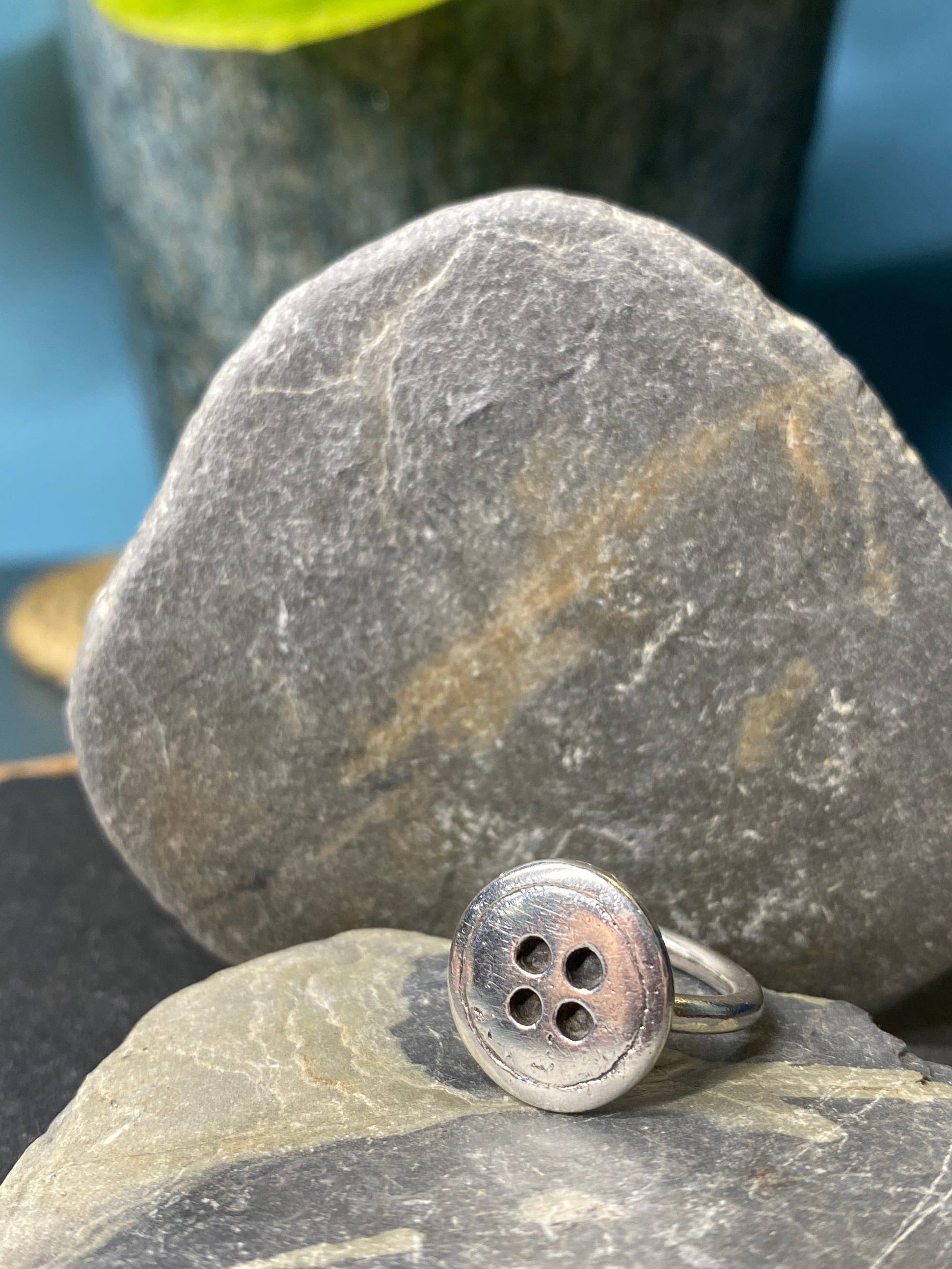 Silver ring with four holes on a stone surface with green leaves in the background. 
Inspired by the Borrowers novel